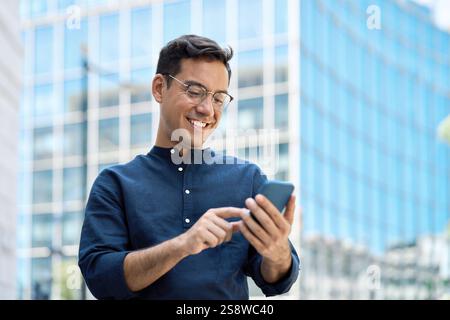 Happy young Hispanic business man using mobile cell phone standing in city. Stock Photo