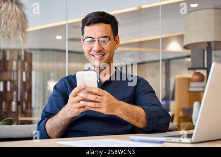 Smiling busy Hispanic business man sitting in office holding mobile phone. Stock Photo