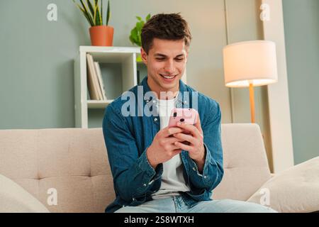 Joyful Guy Browsing with His Cellphone While Relaxing on a Comfy Sofa Surrounded by a Bright Living Room. Young Man Having Fun Using a Mobile Phone at Home. Teenager Watching his Smartphone. High quality photo Stock Photo
