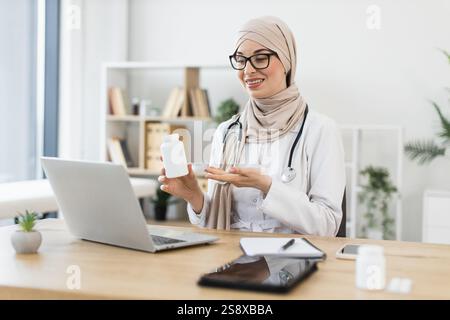 Muslim female doctor in 30s having video call with patient using laptop offering treatment with pills, sitting at desk in medical office, communicating with remote patient online. Stock Photo
