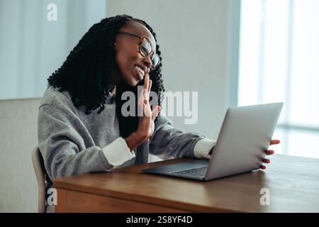 Smiling African woman enthusiastically waves while on a video call, greeting a colleague. She is seated at a desk, using a laptop in a cozy indoor env Stock Photo