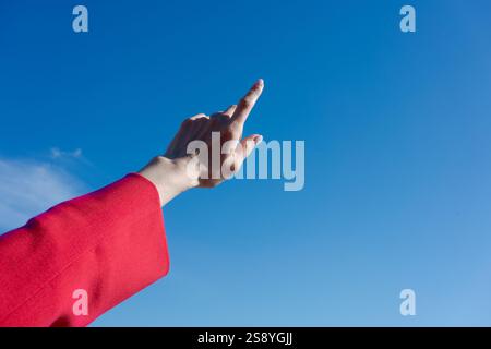 Hand pointing up towards the sky. The sky is blue and clear. The hand is wearing a red jacket Stock Photo