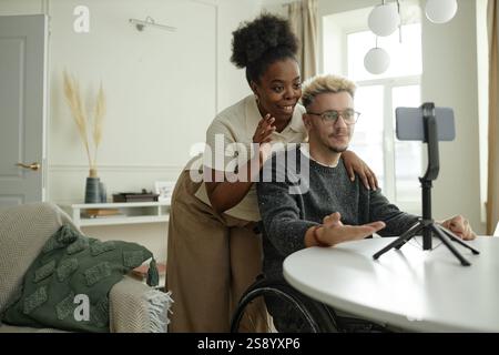 Medium full shot of multiethnic couple of Black woman and man using wheelchair recording video on phone creating content for social media together at home Stock Photo