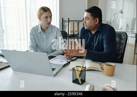 Two colleagues discussing project details in a modern office. Various office supplies and a laptop are visible, enhancing the professional atmosphere Stock Photo
