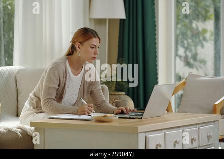 Woman Using Laptop and Taking Notes in Cozy Home Office Stock Photo