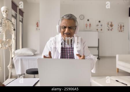 Confident senior doctor wearing headset consulting patient online using notebook Stock Photo