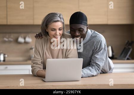 Hugging mature interracial couple using notebook at home kitchen Stock Photo