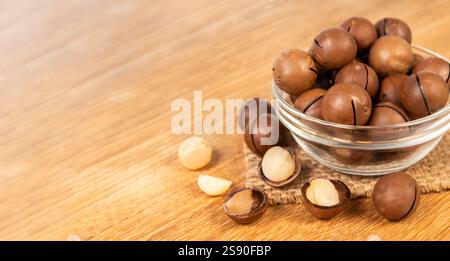 Fresh macadamia nuts displayed in a glass bowl on a wooden table, with a few nuts casually placed around the bowl for emphasis. Stock Photo
