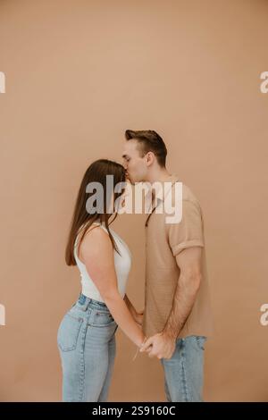 Man kisses woman's forehead, holding hands against a beige backdrop Stock Photo