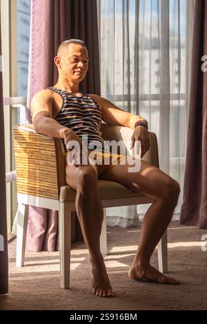 A handsome young Filipino male model in yellow shorts and striped shirt sits on a chair in Cebu City bathed in late afternoon sunlight Stock Photo