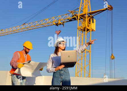 Construction engineers reviewing blueprints and using laptop on site with crane in background, discussing project progress Stock Photo