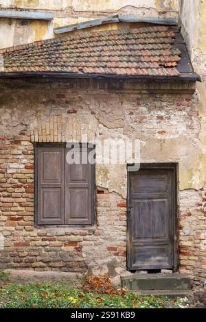 A fragment of an old building with decayed plaster, exposing a brick wall with a wooden door and a window with closed sashes. Stock Photo