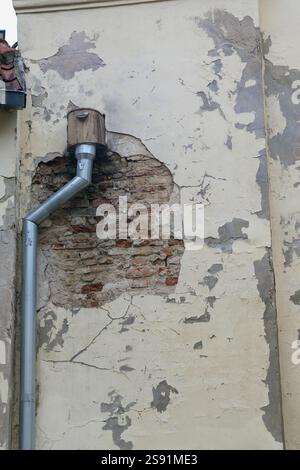 A small hole in the wall of an old building with peeling plaster is visible, in which a birdcage is installed and a rainwater pipe is attached. Stock Photo