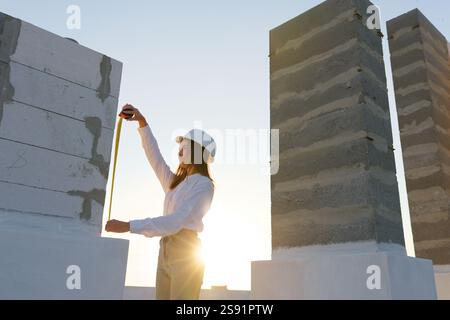 Woman engineer wearing white hard hat using tape measure inspecting construction of concrete block wall at sunset Stock Photo