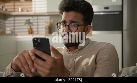 Arabian Indian ethnic man guy use mobile phone on couch browsing mobile internet at home kitchen take break confused read bad news on smartphone male Stock Photo