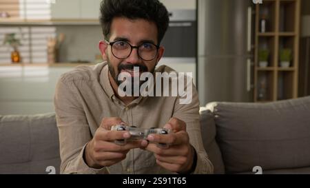 Arabian Indian man guy play video game at home kitchen on sofa male concentrated addicted gamer use console with gaming joystick remote controller Stock Photo