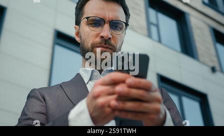 Serious Caucasian entrepreneur adult man businessman CEO business employer office manager outdoors looking at smartphone using mobile phone gadget Stock Photo