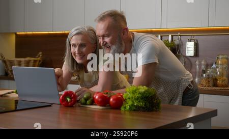 Older Caucasian couple middle-aged spouses family using laptop device at home kitchen choosing food delivery fresh vegetables online shopping natural Stock Photo