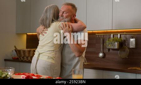 Happy bonding Caucasian old couple retire mature family woman and man dancing in kitchen hugging embracing celebrate anniversary elderly satisfied Stock Photo