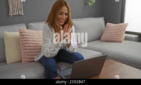 Woman clapping hands while sitting on sofa in living room with laptop, showing joy and enthusiasm in a cozy indoor apartment setting. Stock Photo