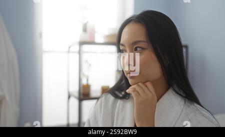 Woman in a spa wearing a robe looks thoughtfully to the side in a serene beauty salon, capturing a sense of relaxation and contemplation in a tranquil Stock Photo