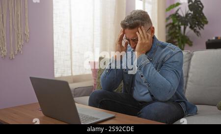 Man sitting indoors looking stressed touching his head while working on laptop in living room at home Stock Photo