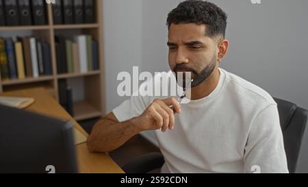 Handsome man thinking in office while holding pen, indoor setting with bookshelves in background and serious expression on face Stock Photo