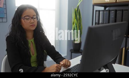 Asian woman wearing glasses working at a computer in an office setting, displaying a focused expression, with a green plant in the background and file Stock Photo