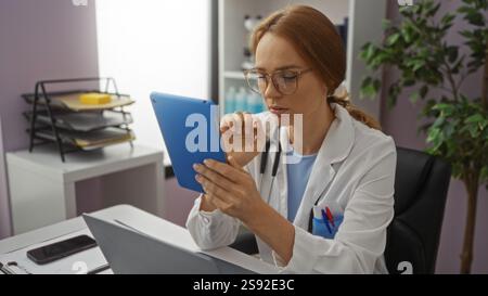 Woman doctor using tablet in modern hospital office, focused on screen, wearing glasses, surrounded by office equipment, displaying concentration and Stock Photo