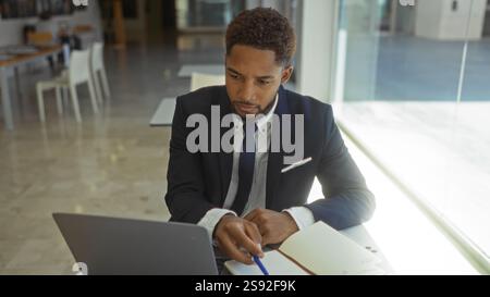 Young man working on laptop in modern office wearing suit writing in notebook focused and concentrated Stock Photo