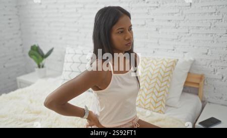 Young woman with back pain sitting on bed in bedroom indoors Stock Photo