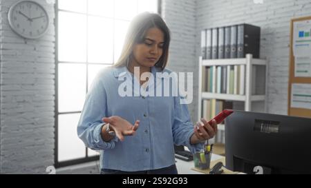 Young woman with brunette hair looking at her phone with a puzzled expression in an office setting, surrounded by shelves, binders, and a clock on the Stock Photo