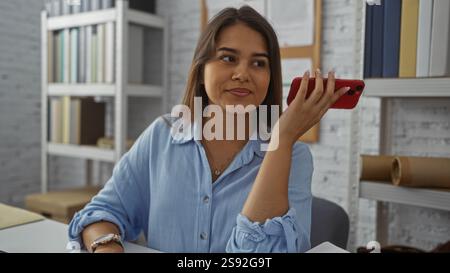 Hispanic woman listening to a voice message on her phone while sitting in an office filled with bookshelves, demonstrating modern workplace communicat Stock Photo
