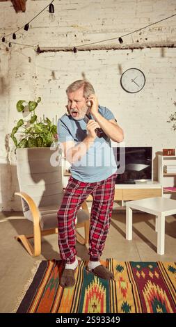Emotional senior man in pajamas listening to music on headphone, singing in tv controller and dancing in stylish light living room at home Stock Photo