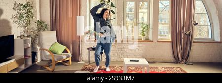 Young man in casual stylish clothes, oversized hoodie and pants, listening to music in headphone and joyfully dancing at home in living room Stock Photo