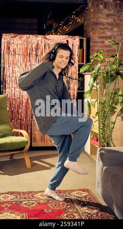 Young man in casual clothes, oversized hoodie and pants, listening to music in headphone and dancing on red carpet at home in living room Stock Photo