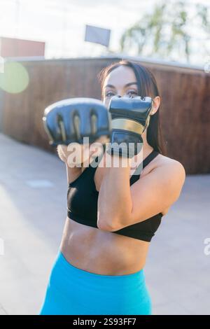 A woman in a black tank top and blue pants is boxing. She is wearing gloves and has her hands up in the air Stock Photo