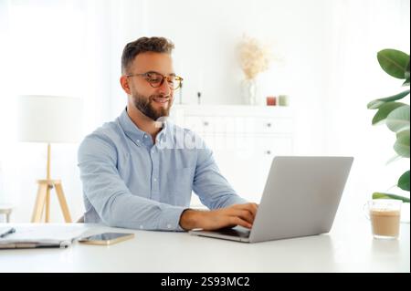 Confident handsome young businessman, company manager or ceo in a blue shirt with glasses, working on his laptop on a new project in a bright and modern office space, surrounded by documents, smile Stock Photo