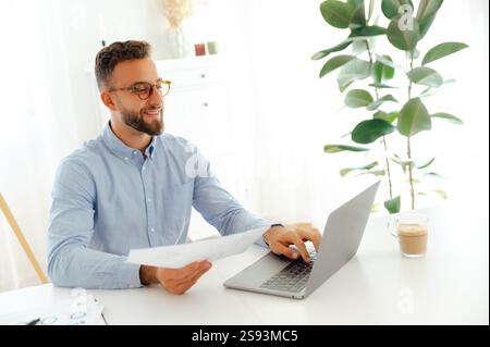 Positive confident businessman works on his laptop while holding papers in a bright and modern office, looking focused as he reviews business charts and plans for success in a project, smile Stock Photo