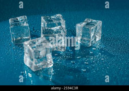 Five transparent ice cubes on a reflective blue surface surrounded by water droplets. A close-up shot highlighting their clarity, freshness, and cool Stock Photo