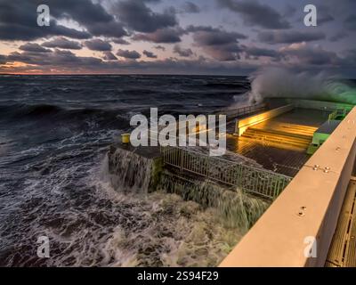 A powerful sea storm with crashing waves covers a pier, surrounded by dramatic clouds during twilight. The image captures nature's force and the beaut Stock Photo