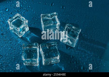 Five transparent ice cubes on a reflective blue surface surrounded by water droplets. A close-up shot highlighting their clarity, freshness, and cool Stock Photo