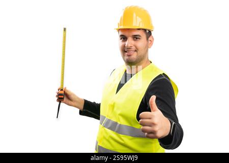Friendly male constructor wearing yellow safety hardhat and fluorescent vest holding tape measure showing thumb-up like gesture isolated on white back Stock Photo