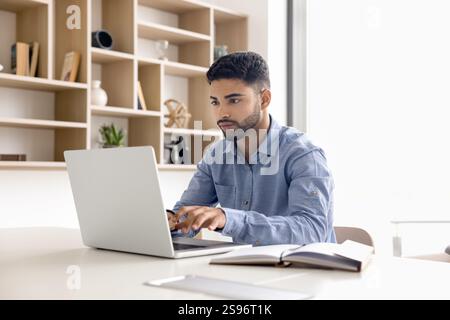 Man sitting at workplace desk, holding pen, typing on laptop Stock Photo