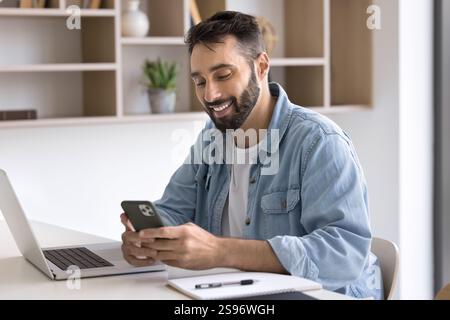 Smiling Latino man sit at workplace desk using smartphone Stock Photo
