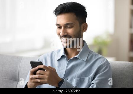 Happy young man sitting on sofa using modern mobile telephone Stock Photo