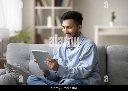 Smiling young man engaged in using pad sitting on couch Stock Photo
