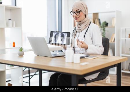 Muslim female doctor having video call with patient showing lung x-ray. Middle-aged woman in medical office using technology for telemedicine consultation. Stock Photo