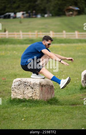 Man Practicing Pistol Squats On A Large Stone Surrounded By Grass And Trees On A Bright And Beautiful Day Stock Photo