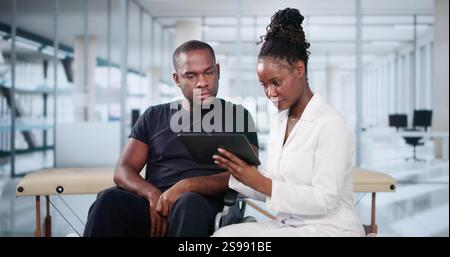 Happy African Doctor With Male Patient In Hospital Using Tablet Stock Photo